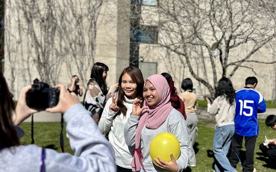 Group of students outdoors, two posing with peace signs, one holding a yellow balloon near a bare tree and building