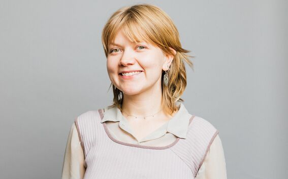 Person with short light hair in layered top and earrings, face blurred, standing against plain grey background