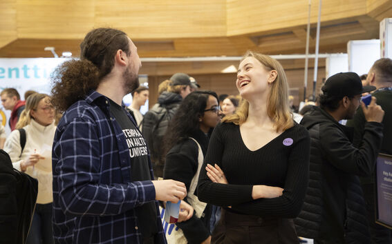 Two people talk at a busy indoor event, standing among a crowd under warm wooden ceiling.