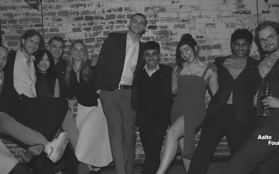 Group of smartly dressed people posing together at a brick‑walled indoor event, some holding drinks.