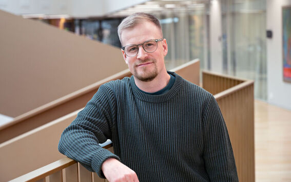 Man with glasses and short beard in dark jumper, leaning on railing in bright modern office
