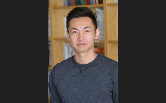 Person with blurred face in a dark grey top standing indoors in front of wooden bookshelves.