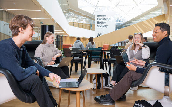 Neljä opiskelijaa istuvat läppärit edessään nojatuoleilla "Better Business Better Society" - kyltin alla.