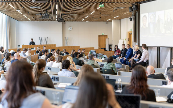 Audience in a modern lecture hall listens to a panel of speakers seated at the front near a large screen.