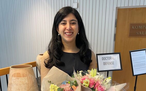 Person in a black dress holding two colourful flower bouquets after a doctoral defence indoors