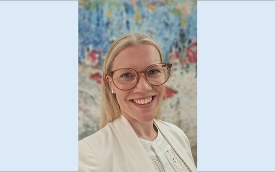 Person in cream blazer and glasses stands indoors before a colourful abstract wall painting