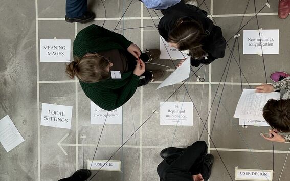 People stand on a gridded floor with labelled papers and strings linking concepts in a workshop activity