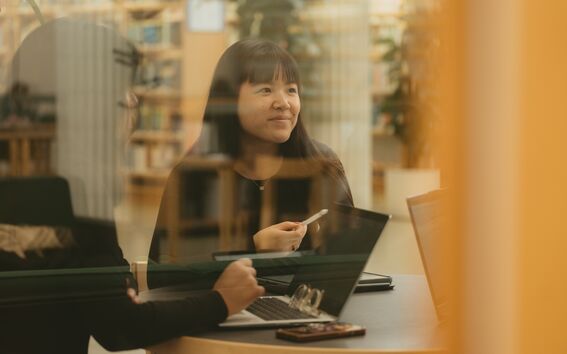 Woman with long dark hair in a library meeting room, holding a pen by laptops, seen through glass.