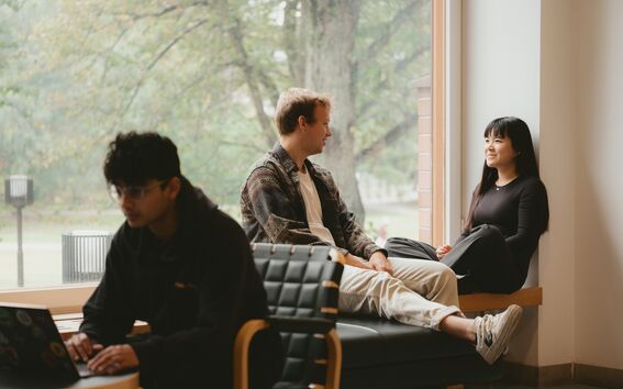 Three students indoors, one on a laptop while two sit by a large window talking, trees visible outside