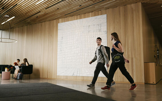 Two students walk through a modern wood-panelled lobby, while two others sit chatting on black sofas