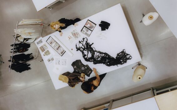 Two people at a large white table designing clothes with sketches and black fabric in a bright studio
