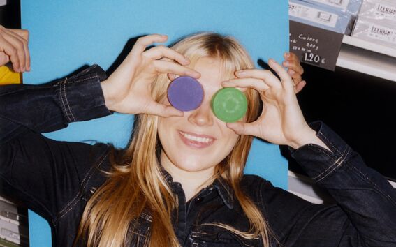 Smiling blonde person holds purple and green discs over her eyes, posing playfully in a colourful shop.