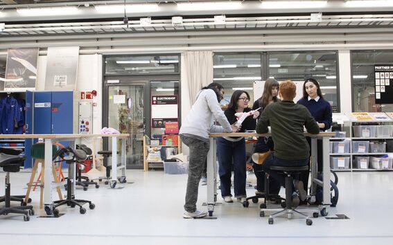 Group of students collaborate around a tall table in a bright workshop with tools and storage shelves.