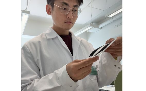 Person in white lab coat and glasses examining a black and white strip in a bright laboratory