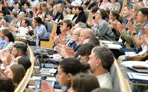 Large, diverse audience in a lecture hall applauding, seated in curved rows with laptops and notebooks