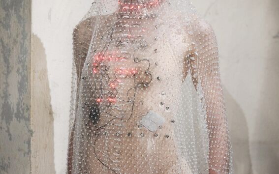 Model in nude bodysuit and sheer beaded veil, wearing visible wires with small red lights on her body