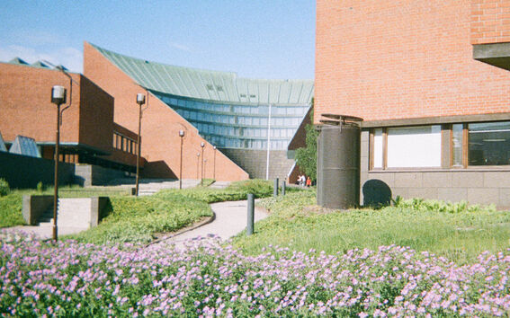 Red brick campus buildings with glass roof and flower beds along a curving path in sunlight