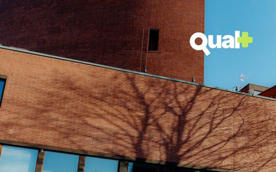 Red brick building with bare tree shadows and blue sky, white text logo “Qual+” in the top right