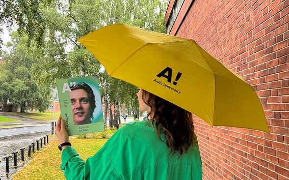 Vihreäpaitainen henkilö keltaisen Aalto-sateenvarjon alla lukee lehteä sateisella kampuksella