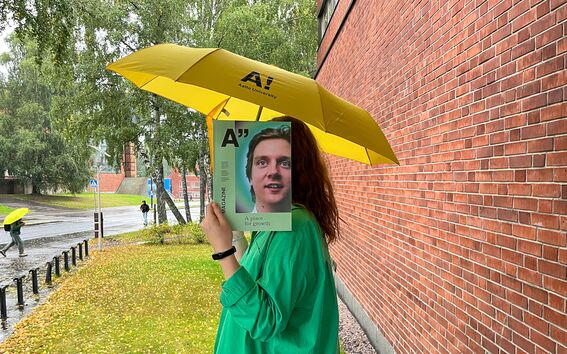Person in green holds magazine over face under yellow Aalto umbrella by wet brick wall and trees