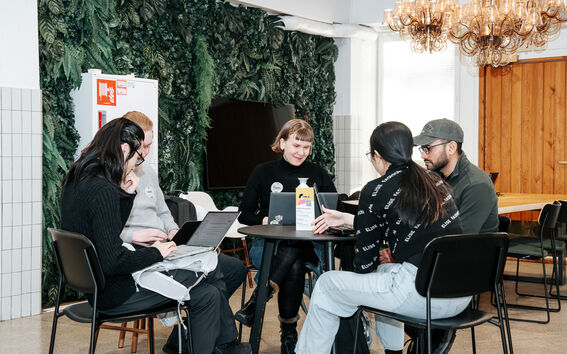 Five people sit around a café table with laptops, working together in a bright room with plants and chandeliers.