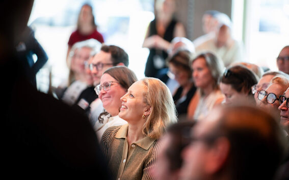 Smiling woman with blond hair in a brown shirt sits among an attentive indoor audience