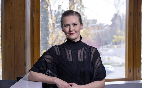 Musta-asuinen nainen nojaa valkoiseen kaiteeseen sisällä, taustalla syksyinen kaupunkimaisema ikkunoista