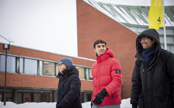Three students in winter coats walk in snow by a red brick campus building with a yellow flag.