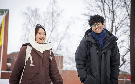 Two smiling students in winter coats walking outside on a snowy campus with bare trees and brick buildings.