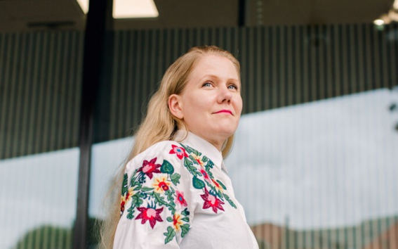 Pitkätukkainen nainen kukkakirjailussa valkoisessa paidassa seisoo ulkona lasiseinän edessä