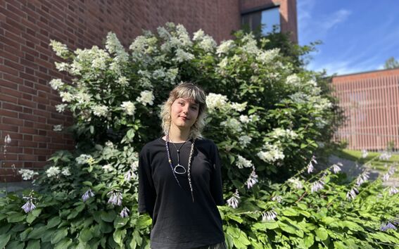 Person in black top stands in a sunny garden with tall white flowering bushes and brick buildings behind.