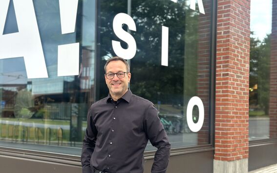 Smiling man in black shirt and jeans stands by large window with white letters and a red brick wall.