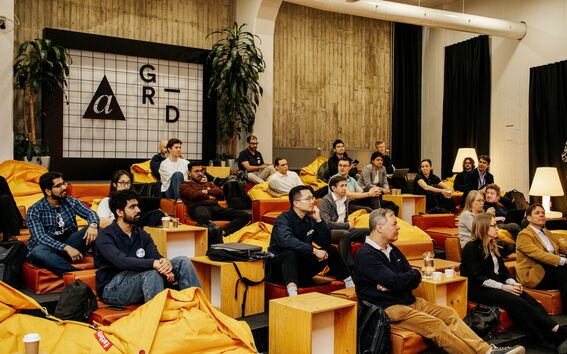 Audience seated on yellow beanbags and wooden steps in a modern hall, listening to a talk.