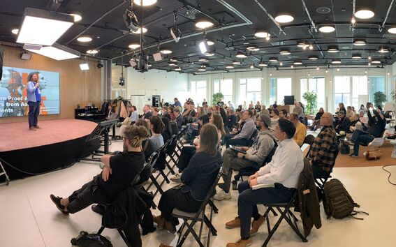 Speaker in blue jacket on stage addressing a full audience in a bright modern conference room