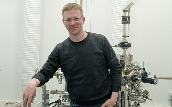 Silmälasipäinen mies tummassa paidassa seisoo kirkkaassa laboratoriossa suuren metallilaitteen vieressä