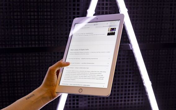 Hand holding a white tablet with text on screen, in front of a dark grid wall and bright light strips