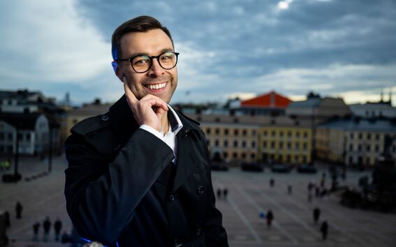 Hymyilevä mies tummassa takissa ja silmälaseissa seisoo kauppatorilla värikkäiden talojen ja pilvien edessä.