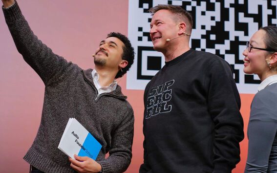 Three people on stage taking a selfie in front of a large black-and-white QR code.