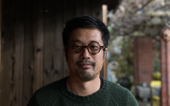Man with round glasses in a dark green jumper, standing under a wooden roof near a garden patio