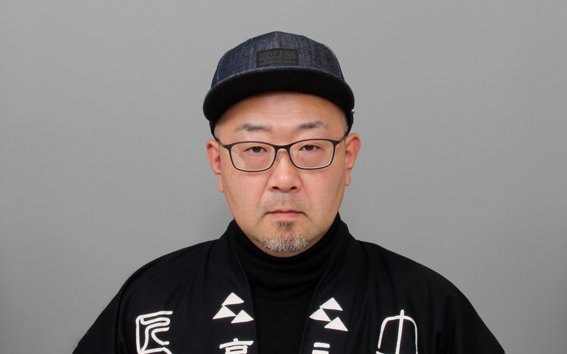 Man in glasses and dark cap wearing black robe with white Japanese text, against plain grey background.