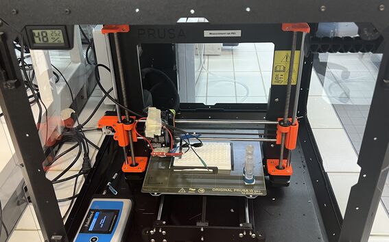 Enclosed Prusa 3D printer with orange parts, wiring and lab tubes on the print bed in a tiled room