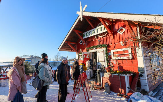 People queue at a small red wooden café in snowy winter sunshine