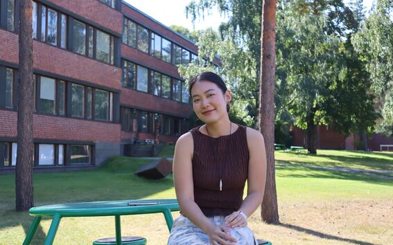 Smiling young woman on a green bench in a sunny campus park with brick buildings and tall trees.