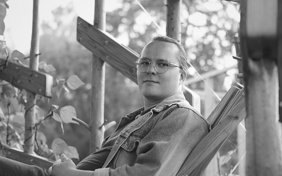 Person in glasses and denim jacket sits relaxed in a wooden garden frame, leaves and soft light around