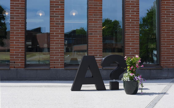 Large black letter A and question mark sculpture beside flower pot outside brick building with tall windows