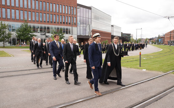 Henkilöitä tummissa puvuissa kävelemässä kampuksella raitiovaunukiskojen yli. Taustalla Väre-rakennus.