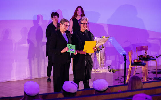 Two women on a purple-lit stage hold green and yellow papers, with flowers and an audience in white caps