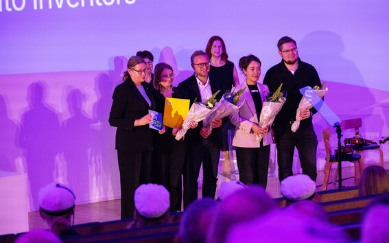 Award ceremony on purple-lit stage, group holding flowers and certificates facing audience in white caps