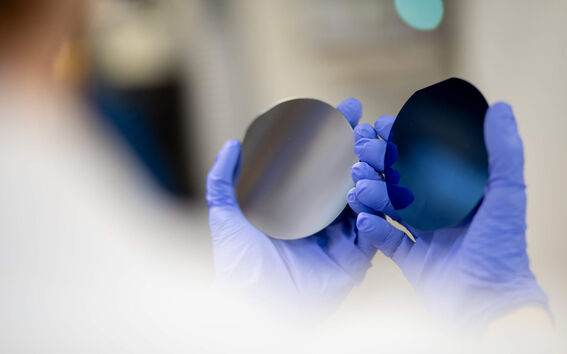 Gloved hands hold two round silicon wafers, one silver and one dark blue, in a bright lab.