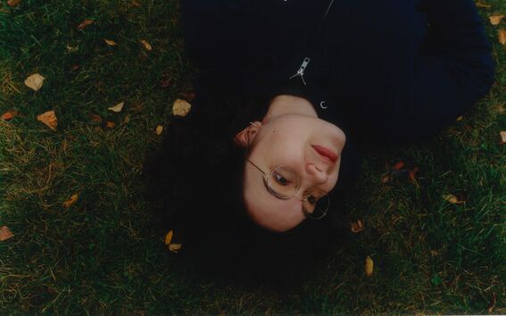 Person in glasses lying on green grass with autumn leaves, wearing a dark Aalto University hoodie